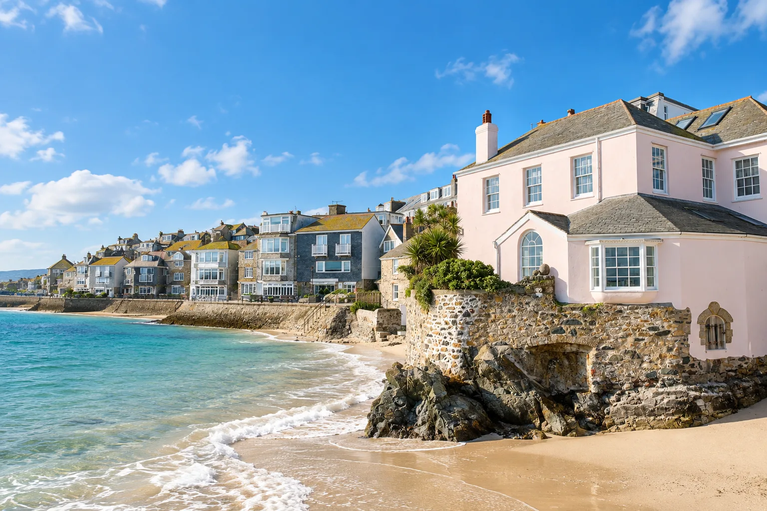 Cottages above the beach at St Ives Bay in Cornwall, with a pink rendered house on the rocks and turquoise water below