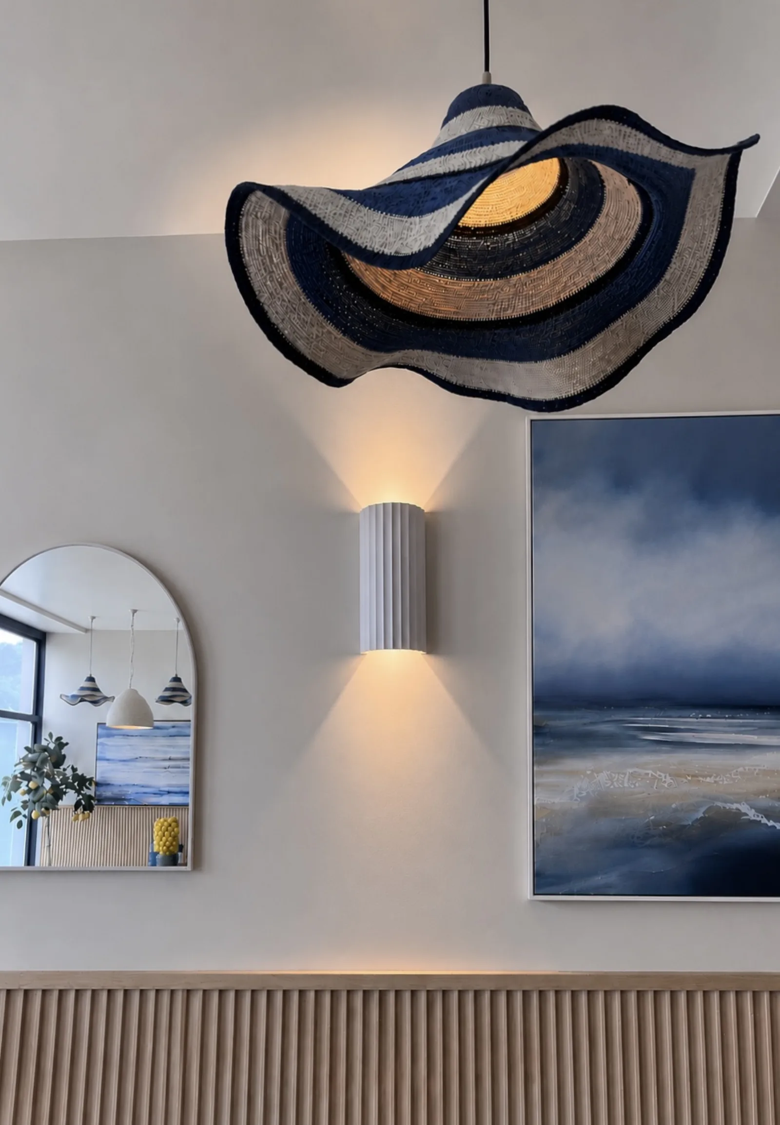 An arched mirror, a ribbed ceramic wall light and a blue and white seascape painting above fluted timber panelling