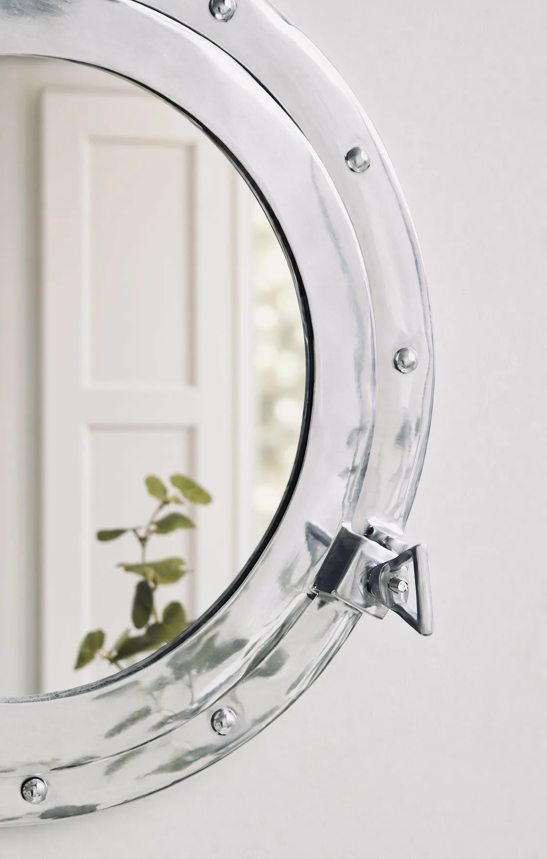 Nautical porthole mirror in polished nickel, reflecting a panelled door and trailing greenery, Appledore, Devon