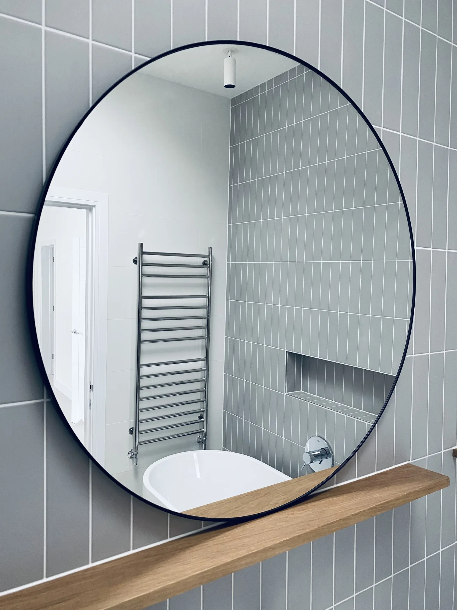 A family bathroom with a round black framed mirror, stacked grey tiling and a solid oak shelf