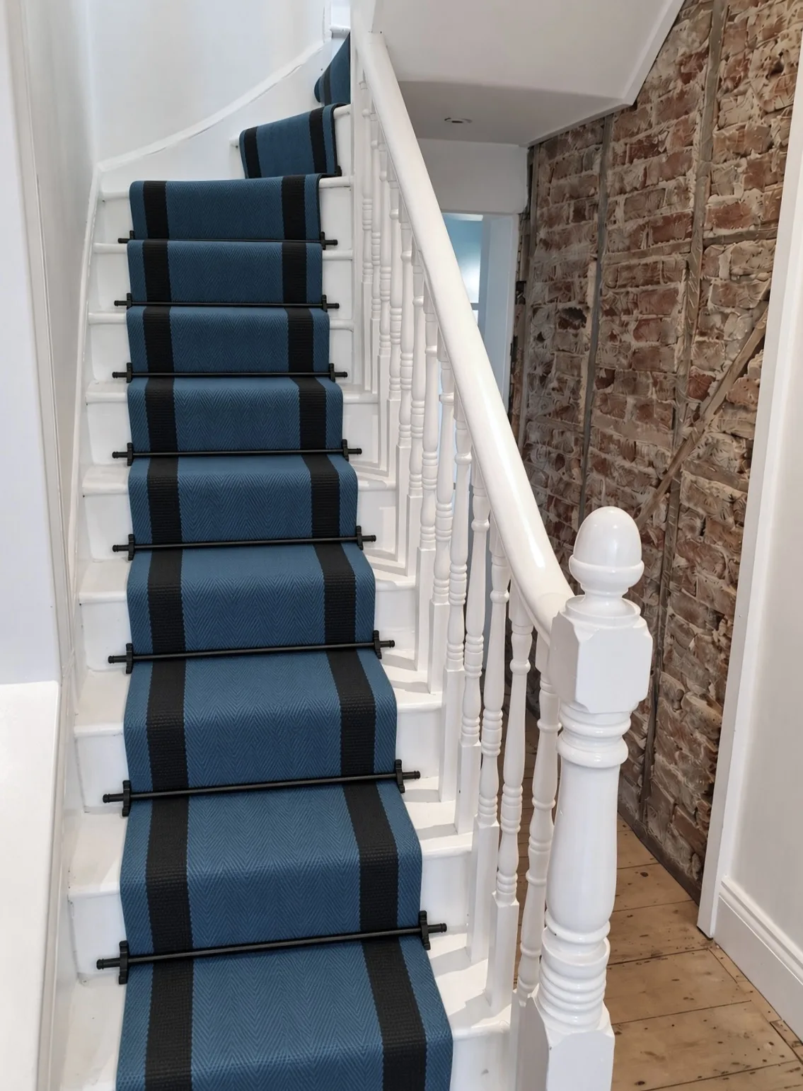 White Victorian staircase with a blue and black striped stair runner on black rods, beside an exposed brick wall, Ethos HQ, London