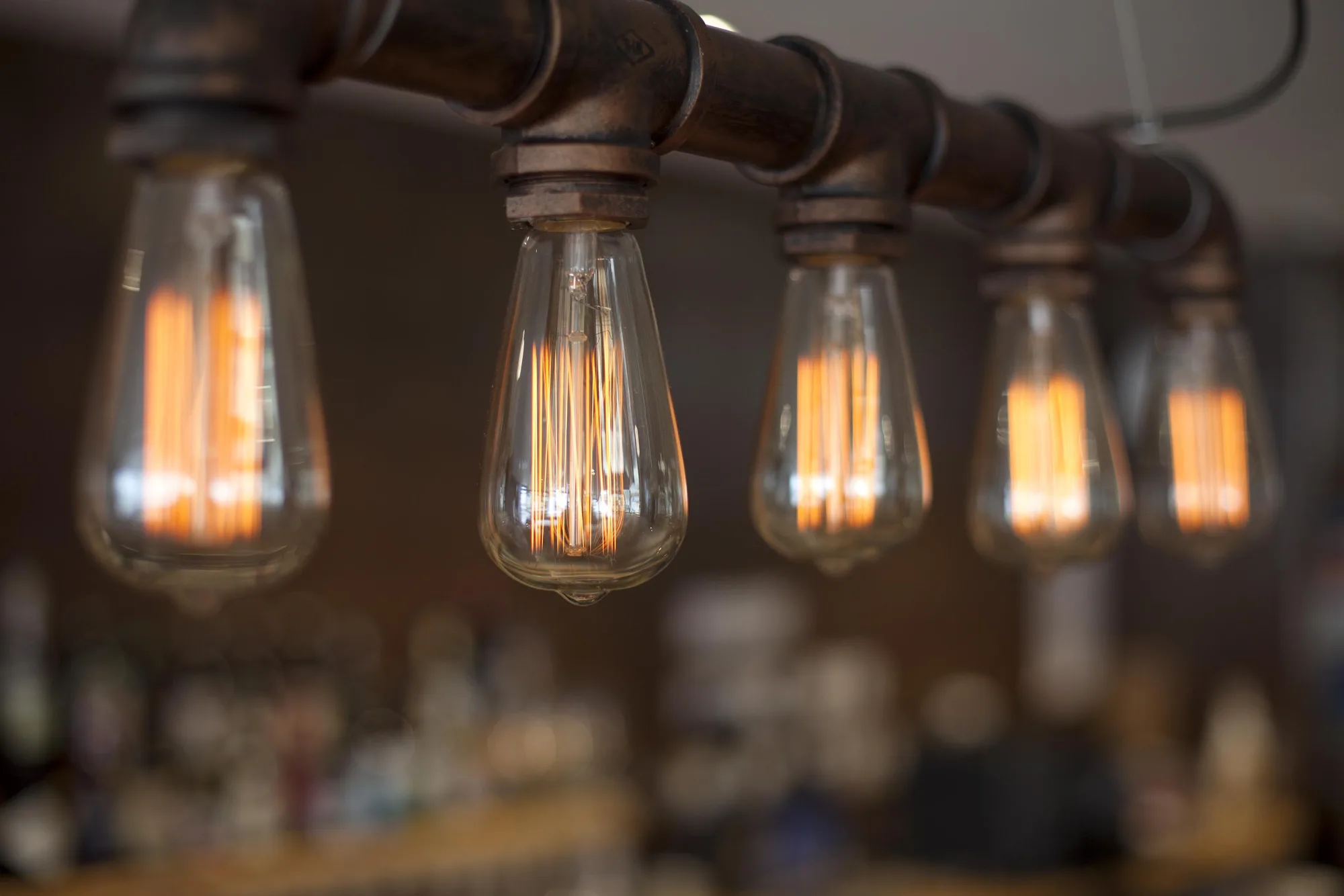 The filament bulbs of the copper pipe pendant seen close, against the exposed brick, at Swell Bar & Kitchen in Truro, Cornwall