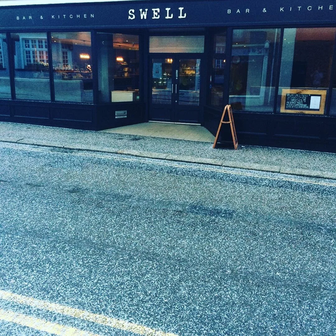 The black timber shopfront and pavement sign seen from across the street, at Swell Bar & Kitchen in Truro, Cornwall