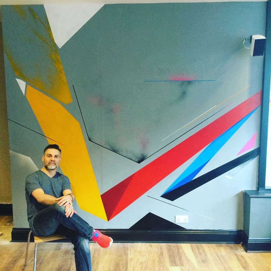 The geometric wall mural in grey, yellow, red and blue, with the artist seated in front of it, at Swell Bar & Kitchen in Truro, Cornwall