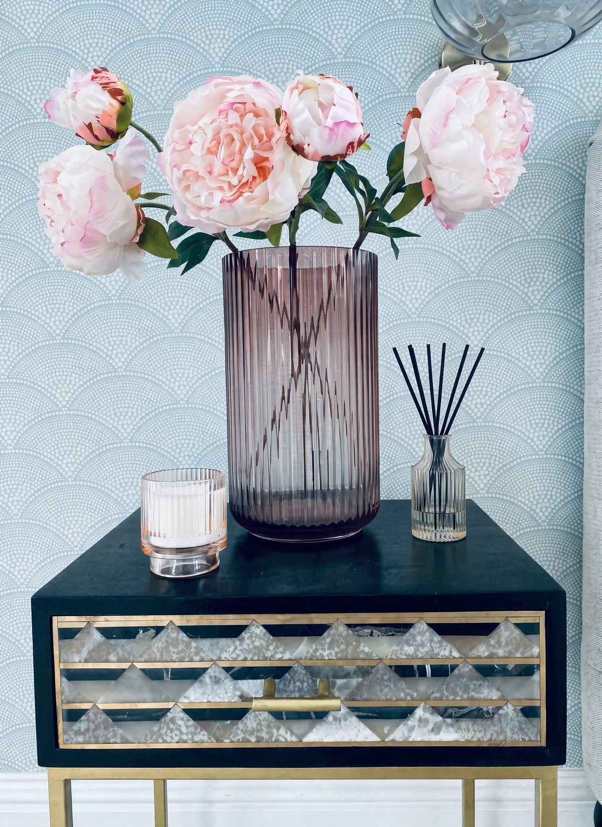 Black mangowood bedside table with a marble and brass inlaid front, styled with peonies and a candle against scallop wallpaper, at an art deco house in Topsham, Devon